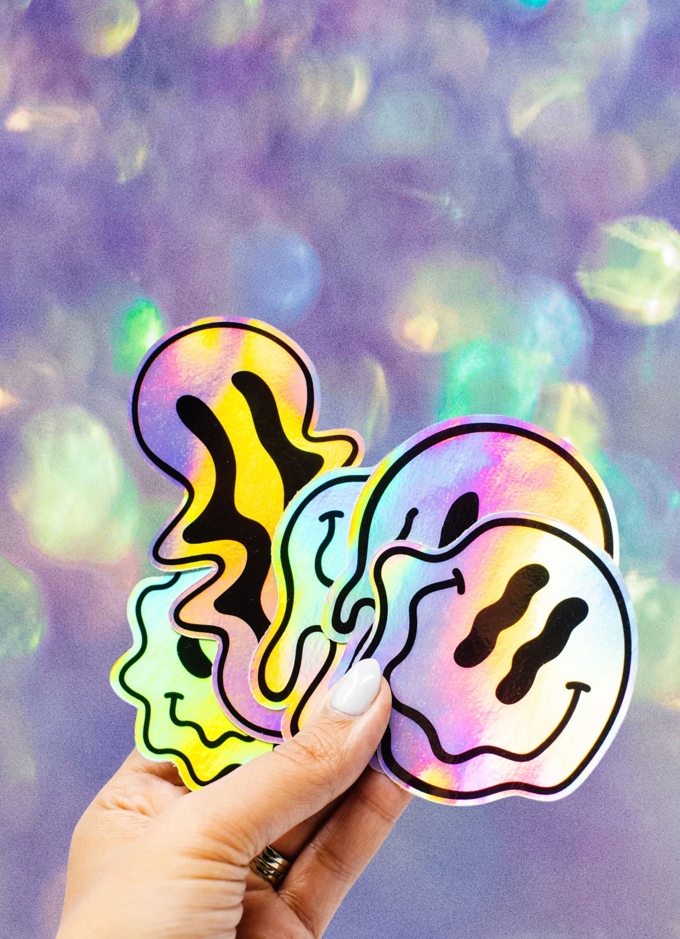 Promotional banner image featuring a hand holding five overlapping holographic smiley-face stickers with black abstract facial details. The stickers reflect rainbow colors against a soft purple and iridescent bokeh-style background. No readable text appears on the image. The scene highlights holographic vinyl material, glossy finish, bold outlines, and vibrant reflective effects used for custom die-cut stickers.