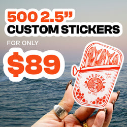 Hand holding a 2.5-inch custom vinyl sticker illustrated like a vintage sardine tin in bold red line art, showing fish packed in oil with floral accents and retro typography. Ocean waves and a soft sunrise backdrop highlight the waterproof aesthetic. On-image text reads 500 2.5” Custom Stickers for only $89, promoting durable die-cut decals.