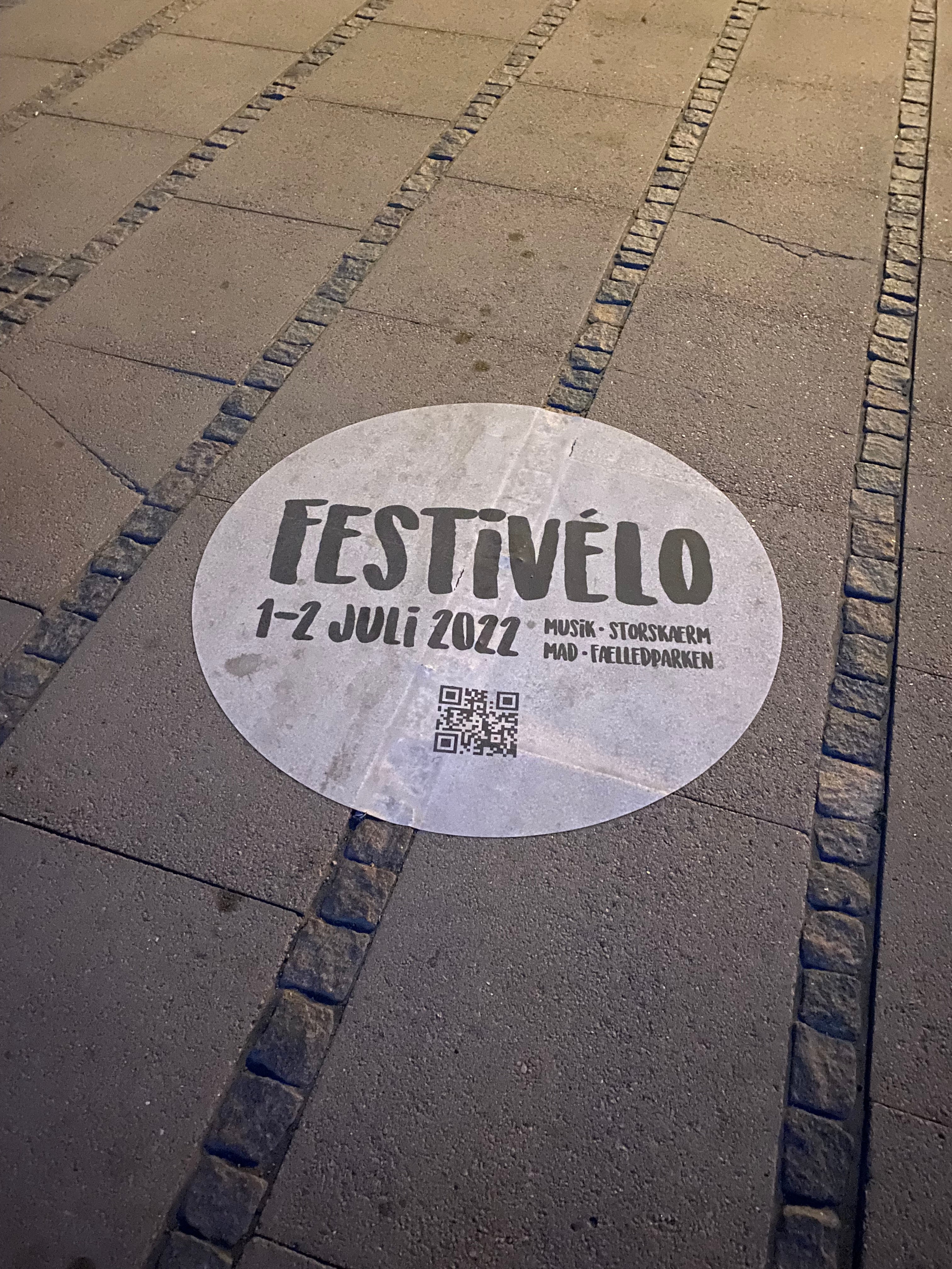 Sidewalk decal advertising a music festival used as temporary promotional signage. black and white sidewalk decal reads. Festivelo 1-2 Juli 2022 Musik. Storskaerm. Mad. Faelledparken