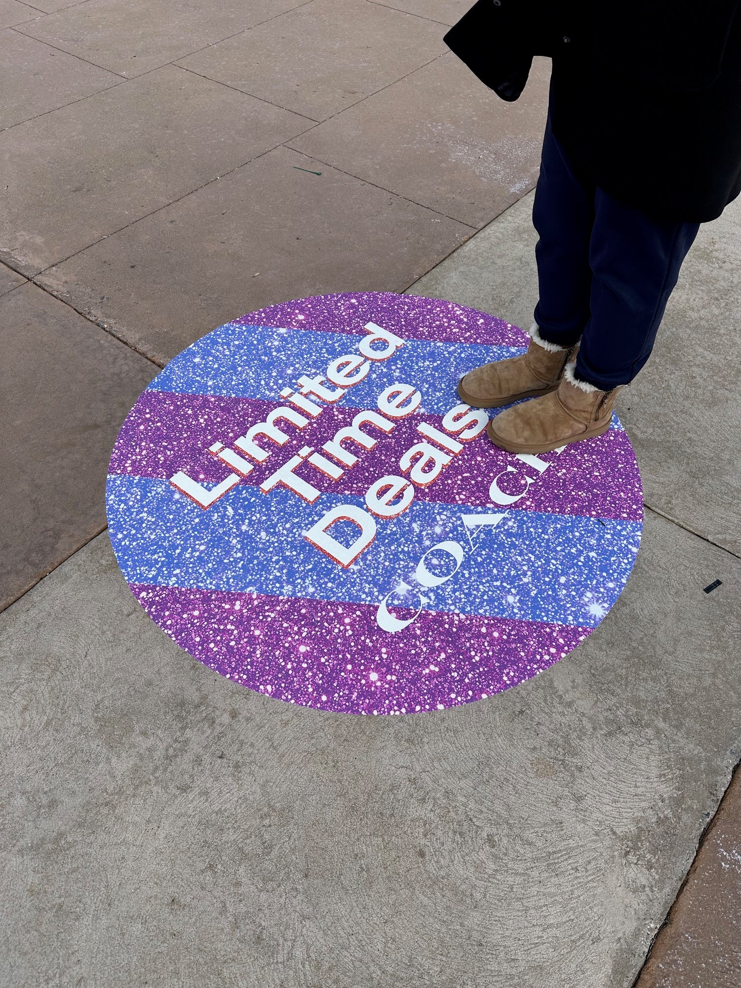 Round, glittery graphic promotional sidewalk decal on a pavement with text. Limite Time Deals. COAC and a foot stepping onto it.