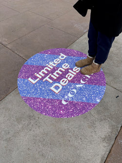 Round, glittery graphic promotional sidewalk decal on a pavement with text. Limite Time Deals. COAC and a foot stepping onto it.