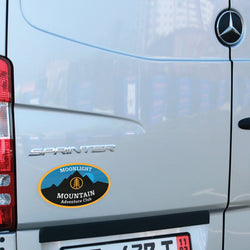 Mercedes-Benz Sprinter van with a 'Moonlight Mountain Adventure Club' sticker on the back. Sticker of mountain and yellow tree icon in yellow circle