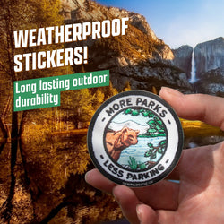 Weatherproof custom vinyl sticker held outdoors in front of a golden autumn mountain landscape. The round die-cut sticker features an illustrated bear beside a lake with pine trees and the text More Parks Less Parking. Bold text on the image reads Weatherproof Stickers! and Long lasting outdoor durability.