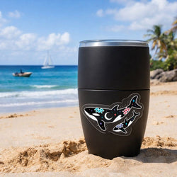 A black insulated tumbler sitting on sandy beach with a clear vinyl sticker of an illustrated orca whale decorated with stars, flowers, and a crescent moon. In the background are ocean waves, a sailboat, a small motorboat, palm trees, and a bright blue sky.
