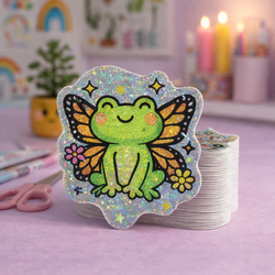 Stack of custom die cut vinyl stickers featuring a smiling green frog with orange and black butterfly wings and small flowers, printed on pixie dust holographic material with dense sparkling glitter. The sticker sits in a pastel desk setting with pink scissors, candles, and soft rainbow wall art blurred in the background.