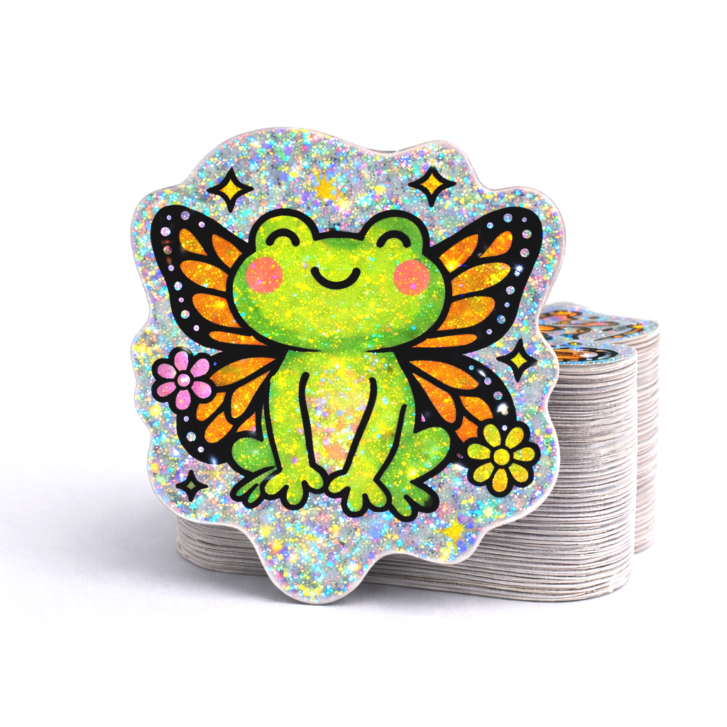 Stack of custom die cut vinyl stickers featuring a smiling green frog with orange and black butterfly wings, pink cheeks, and small flowers. The design is printed on pixie dust holographic material with sparkling glitter flecks throughout the background, creating a bright, shimmering rainbow effect on a light gray surface.