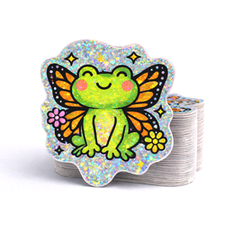 Stack of custom die cut vinyl stickers featuring a smiling green frog with orange and black butterfly wings, pink cheeks, and small flowers. The design is printed on pixie dust holographic material with sparkling glitter flecks throughout the background, creating a bright, shimmering rainbow effect on a light gray surface.