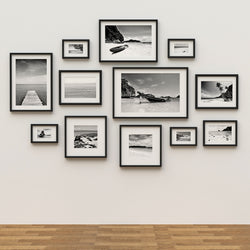 Wall with multiple black and white framed canvas print photographs arranged in a grid pattern. Content is boats and water and beach environments