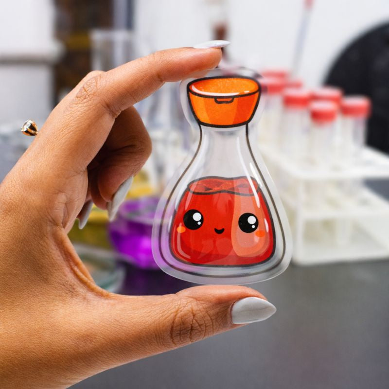 A hand holding a clear vinyl sticker shaped like a small potion bottle with an orange stopper. Inside the bottle is a smiling red liquid character with cartoon eyes and a friendly expression. The background shows a blurred laboratory-style setting with test tubes and colorful liquids.