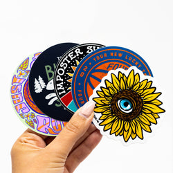 A hand holding four custom vinyl stickers with various designs, including a sunflower, text-based artwork, and geometric shapes. Text Reads/ Your new lucky TS. * 20 * Four. Imposter Syndrome Club. The Bunny Slope. Just Run.