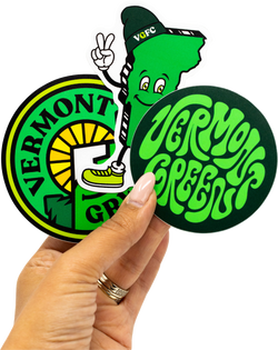 Two Vermont Green soccer club team stickers held by a hand, with one featuring a cartoon character and text. Vermont Green. VGFC.
