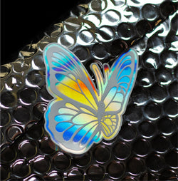 Holographic butterfly sticker with iridescent blue, yellow, and silver wings resting on black bubble wrap packaging. The butterfly design features detailed wing patterns with a reflective metallic finish that shifts color in the light against a dark textured background.