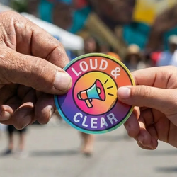 Two hands exchange a colorful round sticker featuring a megaphone graphic and the words 