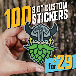 Promotional image advertising ‘100 3.0-inch custom stickers for $29.’ A hand holds a die-cut sticker of a Viking character with a horned helmet, yellow eyebrows, and a green hop-shaped beard. The background shows the textured bark of a tree. The price is highlighted in large yellow text reading ‘$29