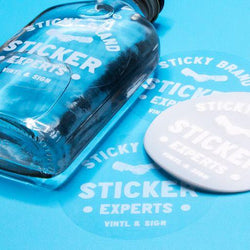Clear circle sticker with white text design label on a glass bottle. Business logo sticker reads. 'Sticky Brand Sticker Expert Vinyl & Sign' blue background