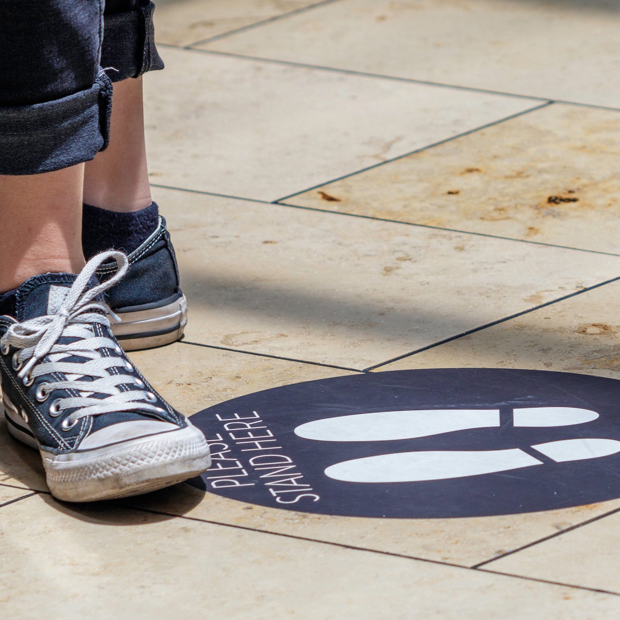 Social distancing sidewalk decal with text. Please stand here and foot print sillhouettes. Stikers on tile imag shows person in blue jeans and sneakers standing next to sticker.
