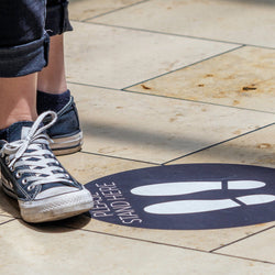Social distancing sidewalk decal with text. Please stand here and foot print sillhouettes. Stikers on tile imag shows person in blue jeans and sneakers standing next to sticker.
