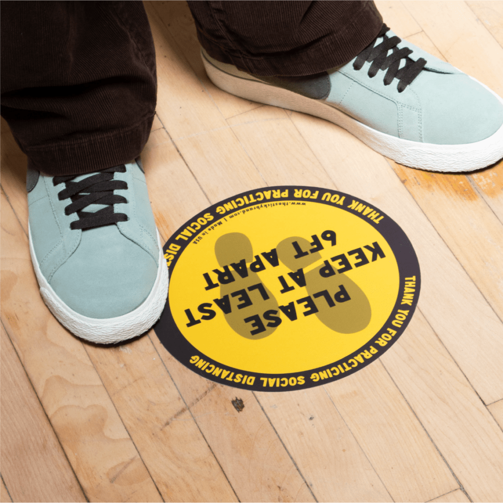 Social distancing safety sticker stuck to hardwood floor and image of person standing next to sticker. The floor sticker graphic reads. Thank you for practicing social distancing. Please keep at least 6ft apart. Sticker is yellow and black