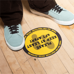 Social distancing safety sticker stuck to hardwood floor and image of person standing next to sticker. The floor sticker graphic reads. Thank you for practicing social distancing. Please keep at least 6ft apart. Sticker is yellow and black