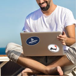 Man on beach with laptop with oval stickers of cape code and ocearch. Red white blue black stickers. Stickers read. Cape Cod, MA. Ocearch Educate, inspire, enable.