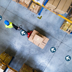 Top view image of man in blue hard hat with neon green vest pulling red dolly with boxes through a warehouse environment following a path created by blue and white arrow floor safety stickers adhered to concrete warehouse floor.