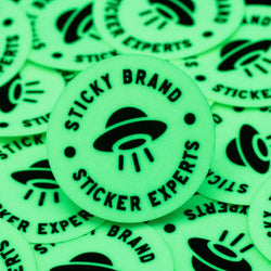 Close-up of glow-in-the-dark round stickers scattered on a surface, each glowing bright neon green with a black UFO and beam icon in the center. Circular text around the UFO reads Sticky Brand Sticker Experts.
